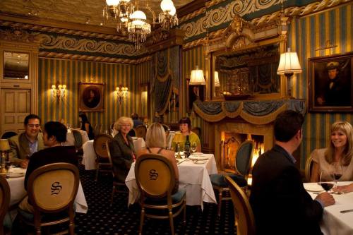 Elegant restaurant interior with diners at tables, ornate decor, and a cozy fireplace. Warm, inviting atmosphere.