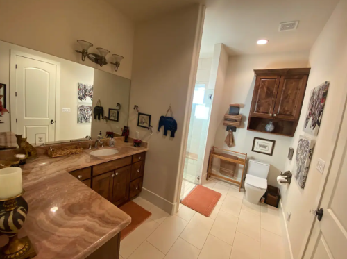 A modern bathroom featuring a double sink vanity, wooden cabinets, and a separate toilet area with light-colored tiles.