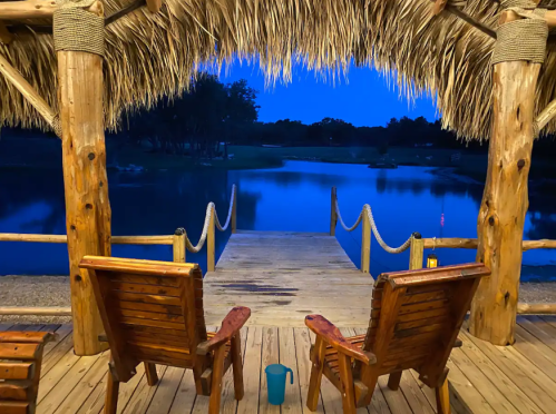 A serene lakeside view at dusk, featuring two wooden chairs on a dock under a thatched roof.