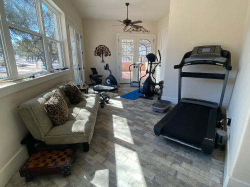 A bright home gym with a treadmill, elliptical, exercise mat, and a cozy sofa against a wall of windows.