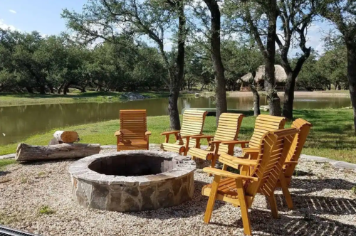 A cozy outdoor fire pit surrounded by wooden chairs, near a calm pond and trees under a clear blue sky.