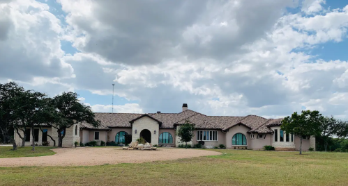 A large, modern house with a stone facade and arched windows, set against a cloudy sky and grassy landscape.