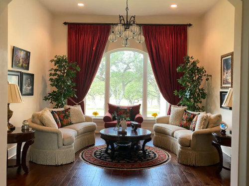 Cozy living room with two sofas, a round table, large windows, and decorative plants, featuring rich red curtains.