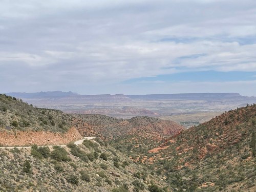A scenic view of rugged hills and valleys under a cloudy sky, showcasing layers of red and green terrain.