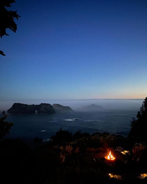 A serene night scene with a campfire, overlooking a misty coastline and distant islands under a starry sky.