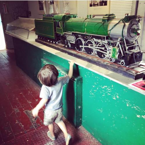 A child explores a model train display in a museum, reaching out to touch a green steam locomotive.