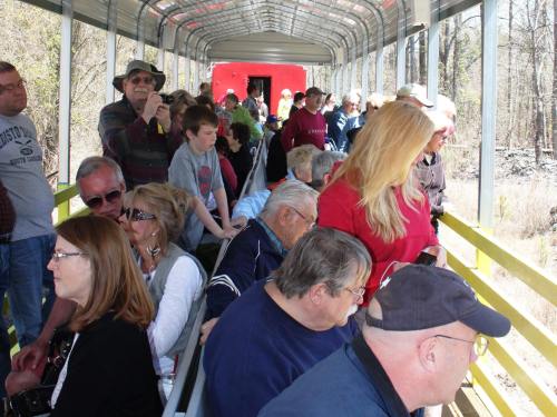 A crowded outdoor tram with passengers of various ages enjoying a scenic ride through a wooded area.