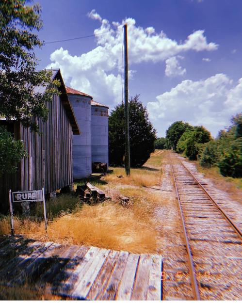 A rustic scene featuring a wooden building, silos, and a railway track under a bright blue sky with fluffy clouds.