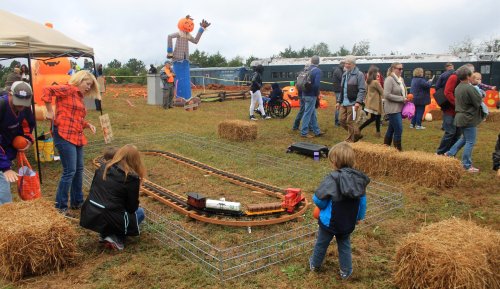 A lively outdoor scene with families enjoying a pumpkin festival, featuring a toy train track and a scarecrow in the background.