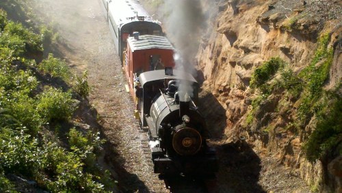 A vintage steam train chugs along a narrow track surrounded by rocky cliffs and greenery. Smoke billows from the engine.