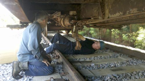 Two men work under a train, one lying on the tracks and the other kneeling beside him, inspecting machinery.