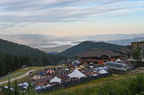 A scenic view of a mountain festival with tents and crowds, overlooking a lake and distant mountains under a blue sky.