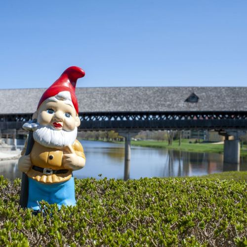 A colorful garden gnome stands in front of a covered bridge and a calm river, surrounded by greenery.