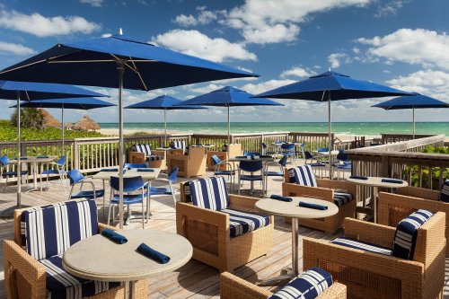 Outdoor dining area with blue umbrellas, striped chairs, and tables overlooking a beach and ocean under a cloudy sky.