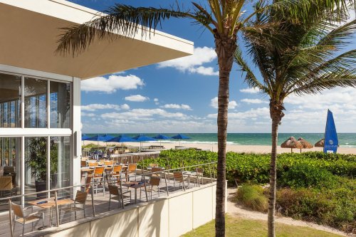 A beachfront restaurant with outdoor seating, palm trees, and umbrellas, overlooking the ocean under a clear blue sky.