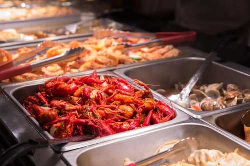 A buffet display featuring various seafood, including red crawfish, clams, and shrimp in metal trays.