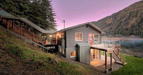 A modern house with a red door, surrounded by trees and mountains, overlooking a calm lake at sunset.