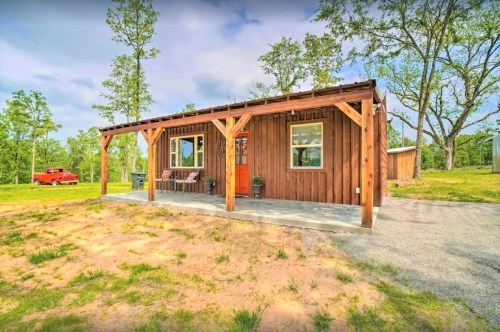 A small wooden cabin with a porch, surrounded by trees and a gravel path, featuring an orange door and a red vehicle nearby.