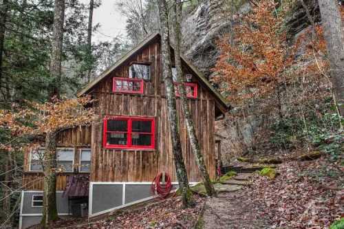 A rustic wooden cabin with red windows, surrounded by trees and autumn foliage, nestled against a rocky hillside.