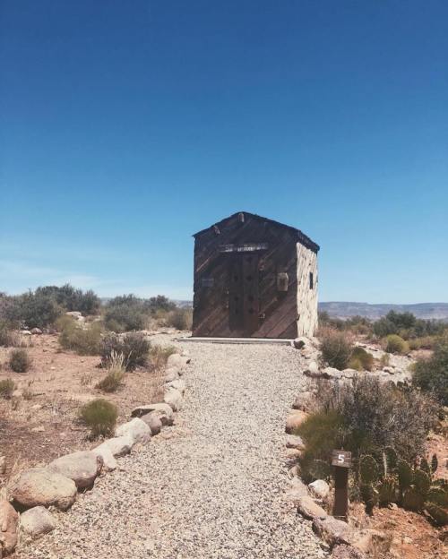A rustic wooden building stands at the end of a gravel path, surrounded by sparse vegetation under a clear blue sky.