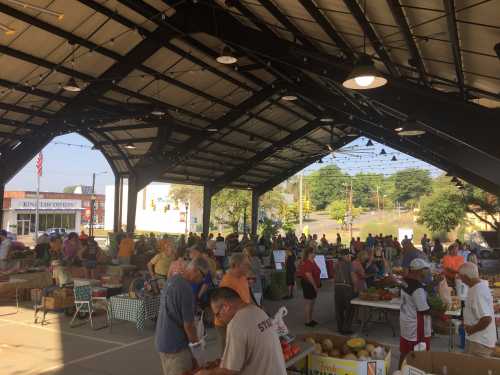 A bustling farmers market under a large pavilion, with many people browsing stalls filled with fresh produce.