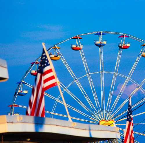 A colorful ferris wheel with seats and an American flag in the foreground, set against a bright blue sky.