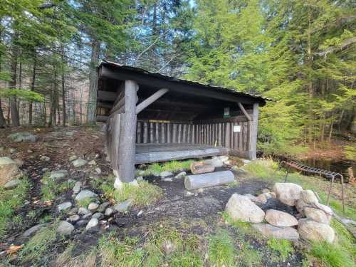 A rustic wooden shelter surrounded by trees, with a stone fire pit and logs nearby.