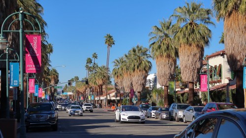 A sunny street lined with palm trees and colorful banners, featuring cars and shops in a vibrant atmosphere.