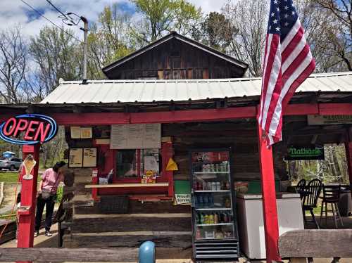 A rustic food stand with an "Open" sign, American flag, and a menu displayed outside, surrounded by trees.