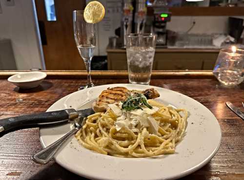 A plate of creamy pasta with grilled chicken, garnished with herbs, alongside a glass of water and a cocktail.