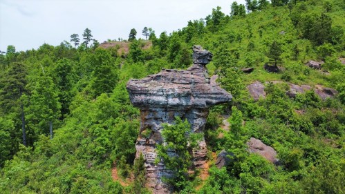 A tall, unique rock formation surrounded by lush green trees and hills under a clear sky.