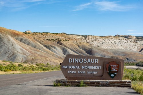 Sign for Dinosaur National Monument at Fossil Bone Quarry, with colorful hills and clear blue sky in the background.