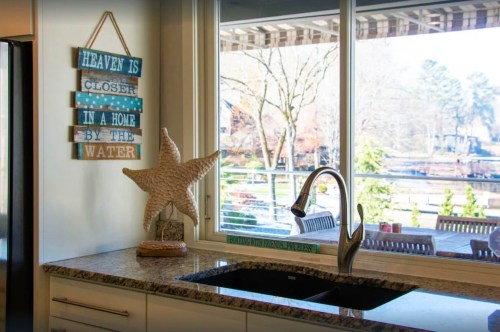 A kitchen sink with a starfish decoration and a sign reading "Heaven is closer in a home by the water," near a large window.
