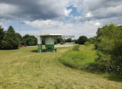 A green observation tower stands in a grassy area, surrounded by trees and under a cloudy sky.
