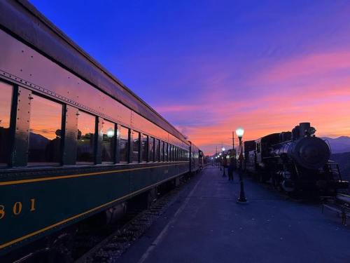 A train station at sunset, featuring a vintage train and colorful sky with reflections on the train's windows.