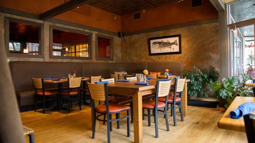 Cozy restaurant interior with wooden tables, chairs, and plants, featuring warm lighting and a welcoming atmosphere.