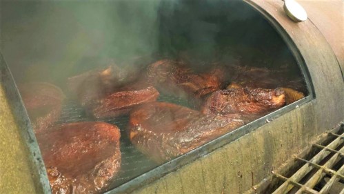 Smoky barbecue meats cooking in a large smoker, with steam rising and a rich, dark color on the surface.
