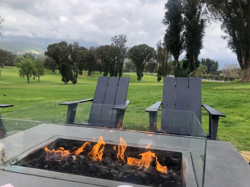 A cozy outdoor scene featuring two chairs by a fire pit, overlooking a green landscape with trees and cloudy skies.