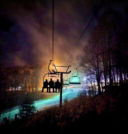 Silhouetted figures on a ski lift at night, surrounded by mist and colorful lights in the background.