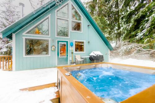 A cozy blue cabin with large windows, surrounded by snow, featuring a hot tub on a wooden deck.