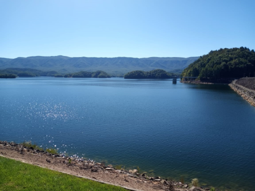 A serene lake surrounded by mountains under a clear blue sky, with sparkling water and a grassy shoreline.