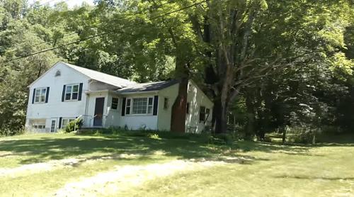 A white single-story house with a green accent, surrounded by trees and a grassy lawn.