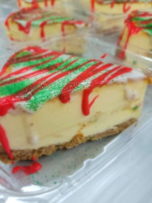 A slice of festive cheesecake with red drizzle and green sprinkles on a graham cracker crust, in a clear container.