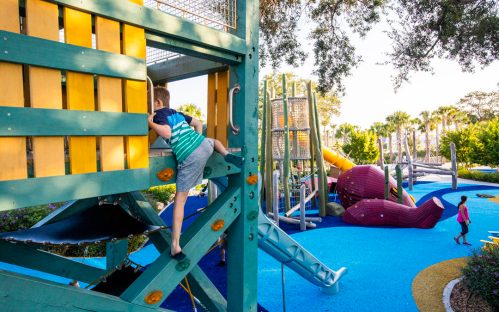 A child climbs a colorful playground structure, with slides and play areas visible in the background.