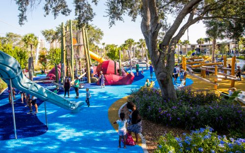 A vibrant playground with slides, climbing structures, and children playing among trees and colorful landscaping.