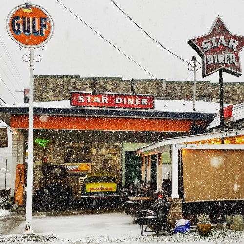 A retro diner with a bright sign, surrounded by snow, featuring a vintage car parked outside.