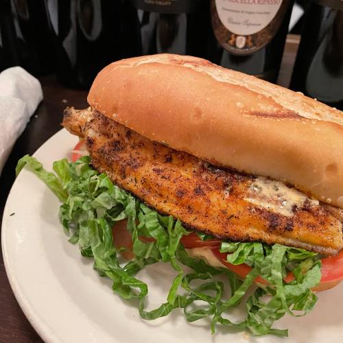 A sandwich with grilled fish, lettuce, and tomato, served on a toasted bun, with bottles in the background.