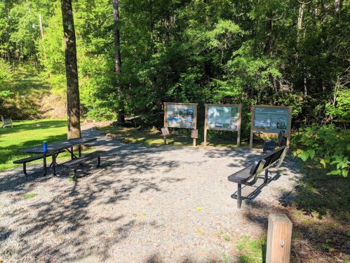 A small clearing in a forest with benches and informational signs surrounded by trees and gravel.