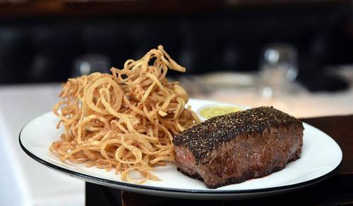 A plate featuring a perfectly cooked steak with a crust, accompanied by crispy curly fries and a side of sauce.