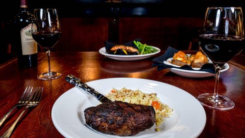 A beautifully plated steak with rice, accompanied by wine glasses and side dishes in a cozy dining setting.